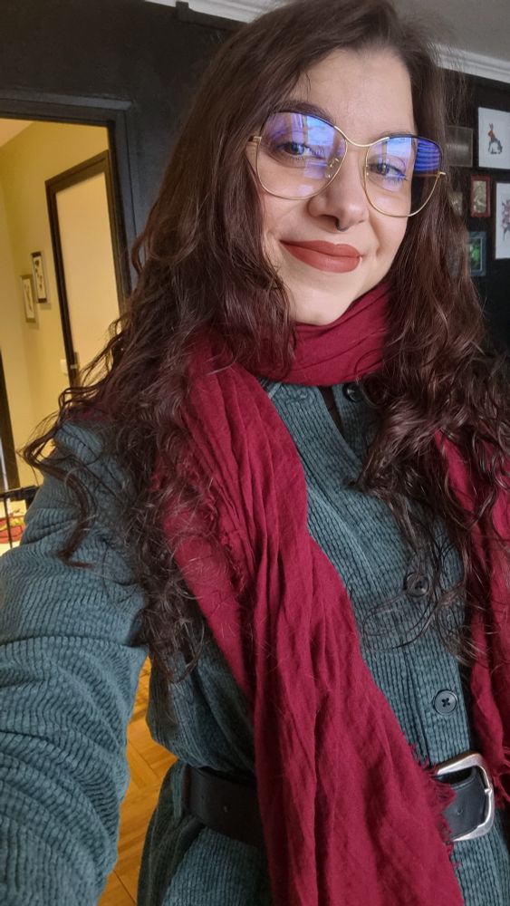 Selfie de ma personne (blanche, cheveux longs auburn ondulés, yeux marrons, grandes lunettes dorées) portant une combinaison en velours côtelé verte ornée d'une épaisse ceinture noire en simili cuir, une écharpe bordeaux légère autour du cou. Je regarde l'objectif en souriant.