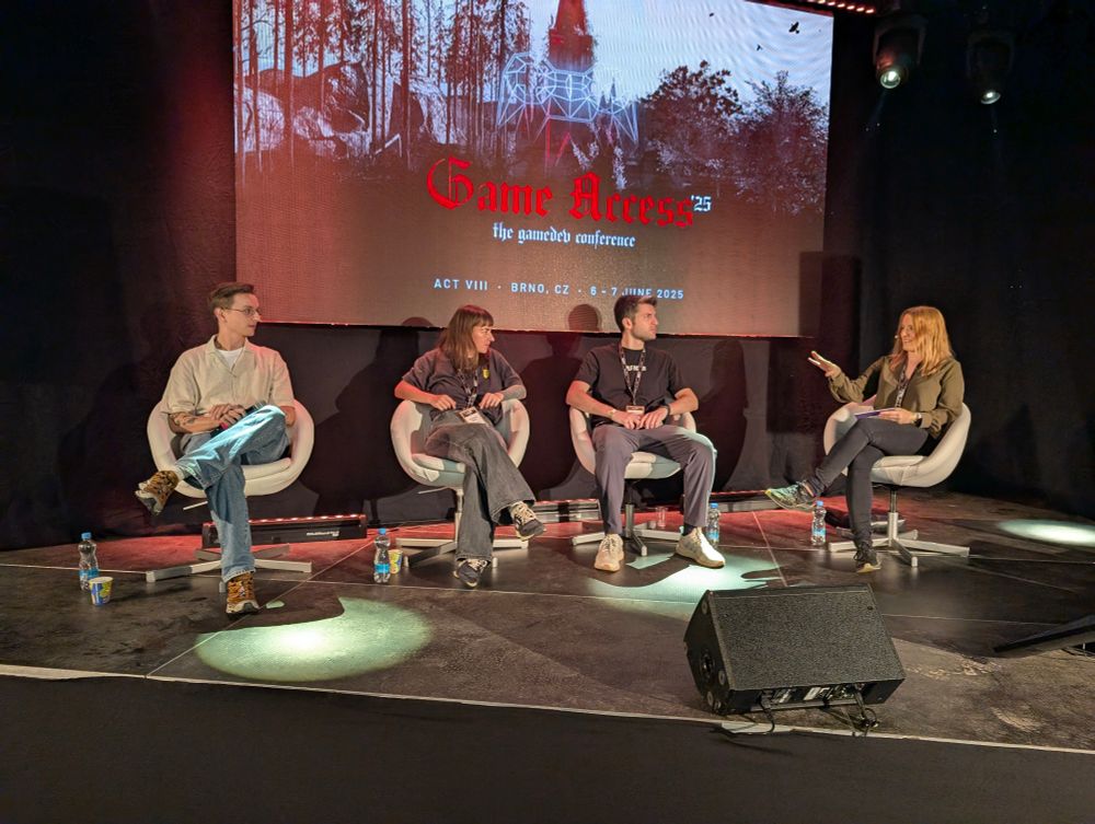 Four people sitting on a stage in a conversation, two men and two women, with one of the women visibly asking a question. The stage has a background with a screen showing the stylized key visual of Game Access