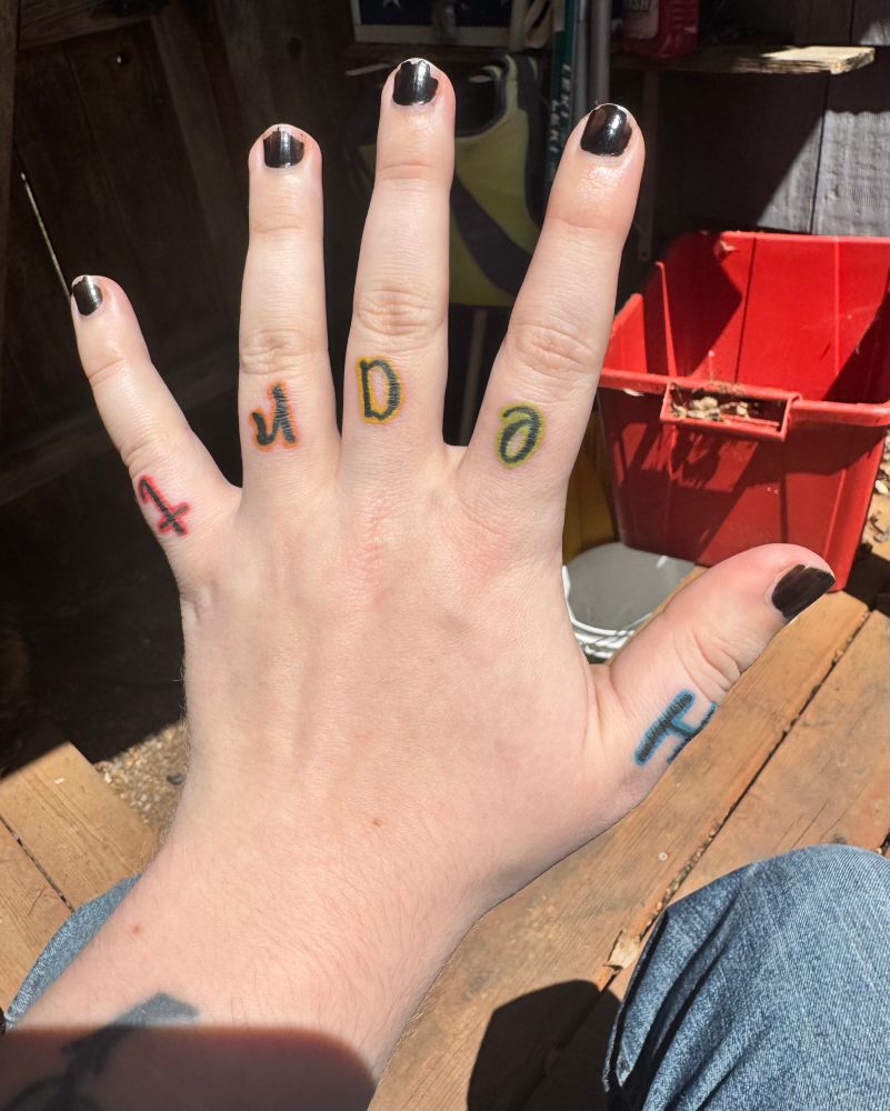 Sun lit photo of honeys left hand with knuckle tattoos that read “Heart” in a swirly font with rainbow outlines
