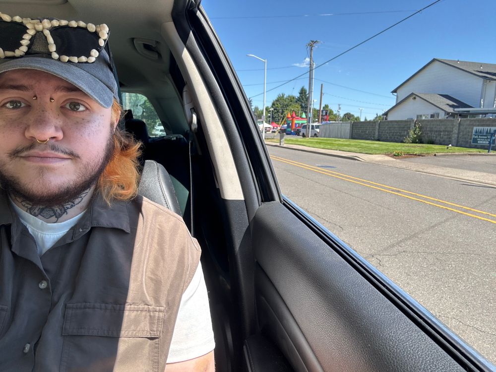 Honey- a white, trans masc, non binary person with a beard- sitting in the drivers seat of his car with the window rolled all the way down. He’s on the left edge of the photo wearing an olive green dress, white tshirt, and a Totoro baseball cap with his teeth sunglasses propped up by the bill of his hat. He has his camera angled to show the brightly colored bouncy castle a block behind him, but you mostly see the road and the apartment complex behind the fair.