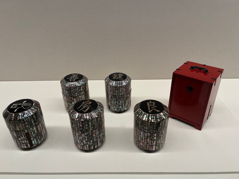 黒田辰秋　螺鈿くずきり容器、岡持ち
螺鈿細工が施された円筒形の小箱が5つ並び、それぞれ蓋に漢字が描かれている。右側には光沢のある赤い漆塗りの箱が置かれている。