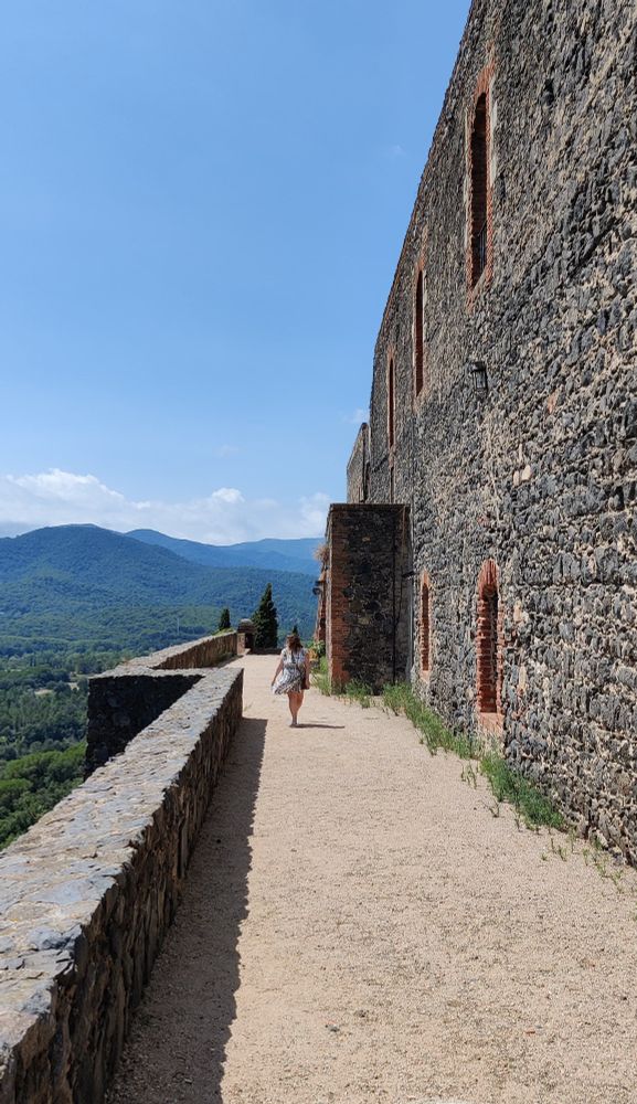 Un muro fortificado muy alto con una persona muy chica y un monte muy azul al fondo.