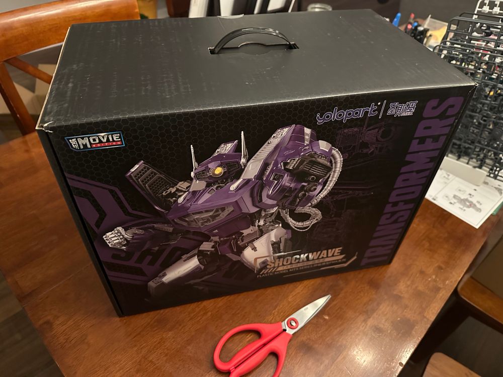 A photo of the box for the Yolopark Shockwave Transformers plastic model kit. It absolutely dwarfs the scissors sitting on the table next to it 