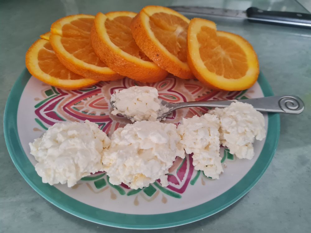 A plate with slices of oranges and clumps of cottage cheese on. There is a silver spoon in the middle and the plate has a greenish blue outer rim with an ornate design in the middle. 