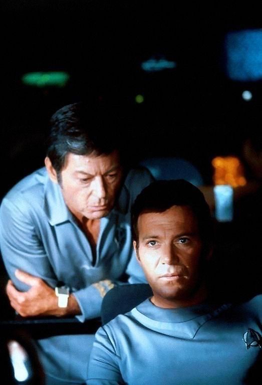 William Shatner’s Jim Kirk sits in the captain’s chair of the USS Enterprise, DeForest Kelley’s Dr. McCoy leaning over his shoulder to speak to him. Both are in the blue-grey uniforms of The Motion Picture. Why Bones’s is cut so low I will never know, but I cannot complain. 