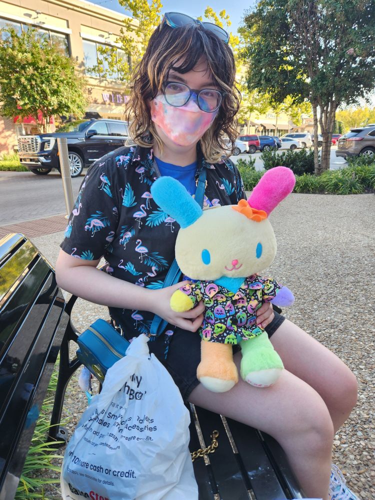 A photo of Luce a pale person with dark hair sitting on a bench with a plush of Usahana from Sanrio, a colorful bunny.