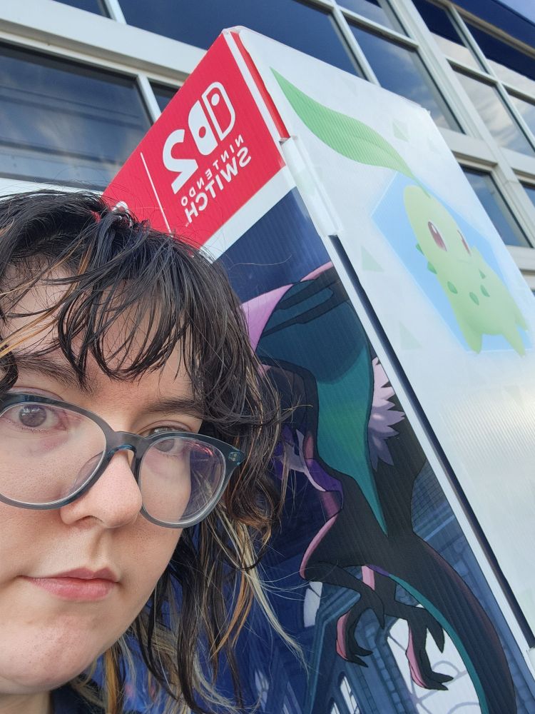 Photo of Luce, a pale person wity medium length dark hair, next to a store display of Pokenon Legends Z-A