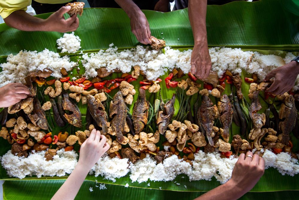 Several hands reach for fish, tomatoes, and other foods arranged on a green leaf on a table.