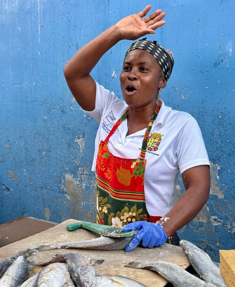 A woman wearing a white shirt, black headband, and red apron raises her hand and smiles as she sings while processing fish. She stands in front of a blue wall and holds a tool to remove scales from fish in her gloved hand. There are silver fish on a cutting board in the foreground.