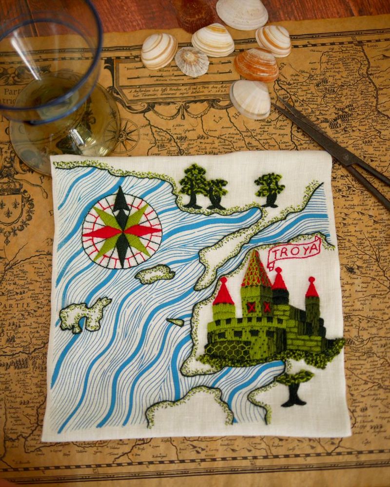 Une carte marine brodée de la côte Turque au niveau la ville de Troie. La toile de lin blanc est posée sur une table sur une carte ancienne avec quelques coquillages et un verre à pied. La mer, représentée par des vagues bleues est imprimée. La côte, ainsi qu'une rose des vents et une cité fortifiée sont brodées en différentes nuances de vert et du rouge. Le long de la côte, des petits points sont brodés et quelques arbres au feuillage chaotique.