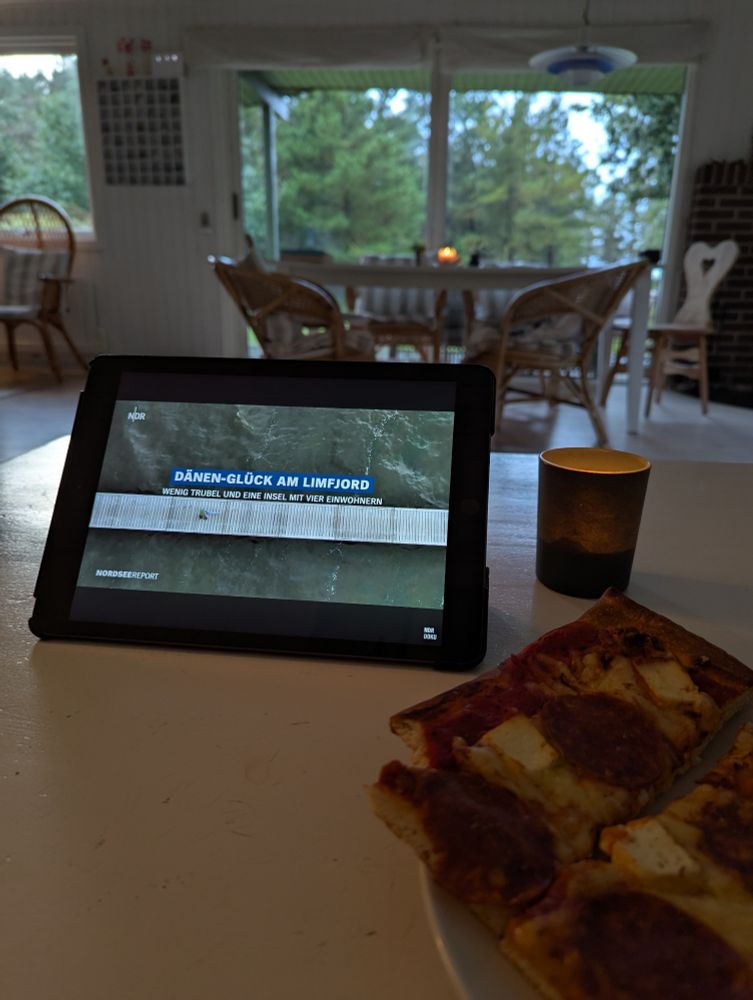 Ein iPad aud einem Tisch, davor eine Pizza mit veganer Salami und Hirtenkäse. Auf dem iPad läuft der Nordseereport "Dänen-Gluck am Limfjord"