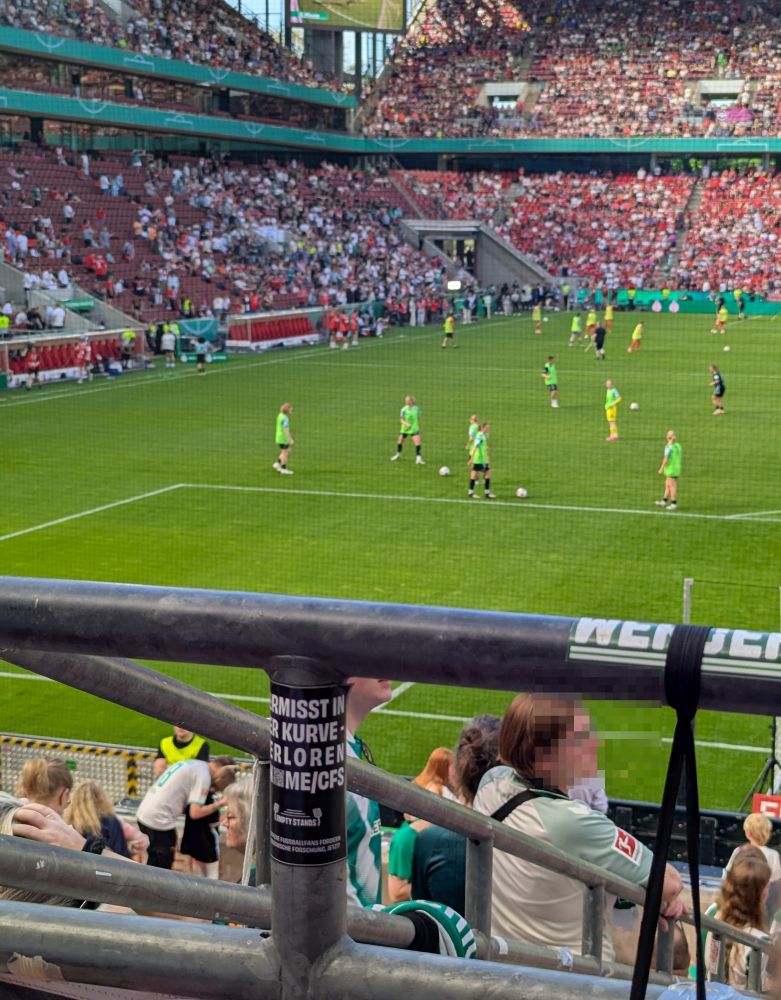 Ein Empty Stands Aufkleber "Vermisst in der Kurve, verloren an ME/CFS" an einem Geländer im Müngersdorfer Stadion im Werder-Block, auf dem Feld wärmen sich Spielerinnen von Werder und Bayern auf