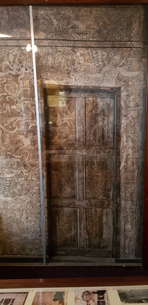 Fresco of angels, demons and winged beasts, surrounding and continuing onto a historical wooden door. Covered by glass to protect it.