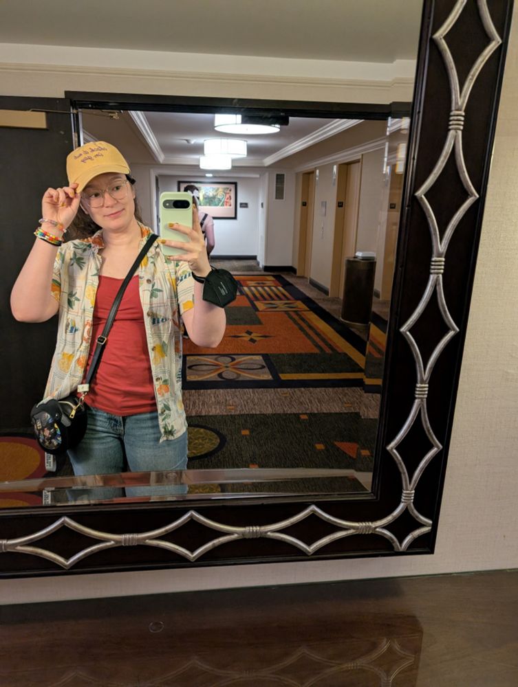 A mirror selfie with a beach-themed button-up and a goldenrod hat that says "check it out, I'm gay" 