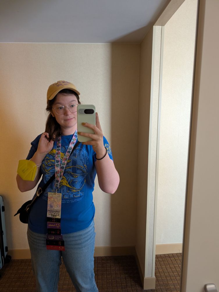 a mirror selfie with an AGDQ2024 volunteer shirt in blue, a GDQ mascots lanyard, yellow mask on wrist and a yellow hat that says "check it out, I'm gay". 