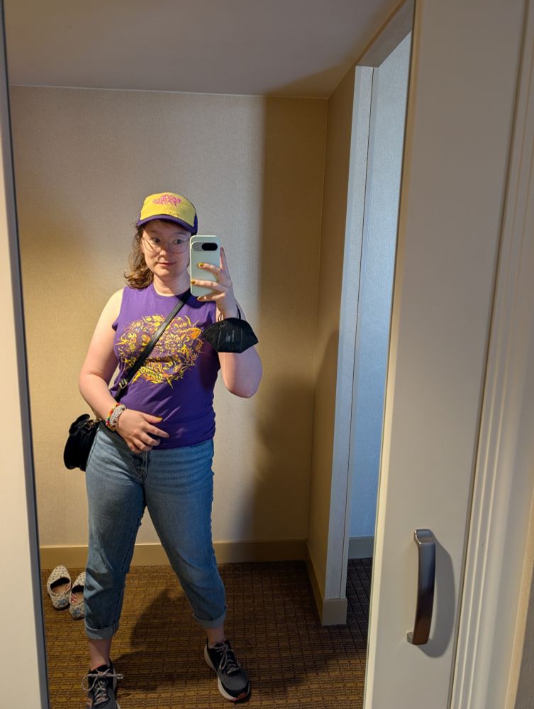 A mirror selfie. They are wearing a Flame Fatales 2022 tee-shirt and a homemade Frame Fatales ballcap. They have friendship bracelets on one wrist and a KN95 mask on the other. 