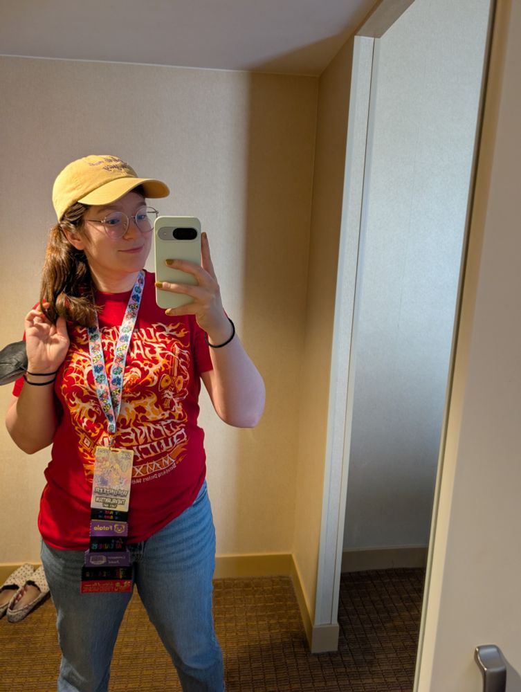 Mirror selfie with a red volunteer shirt for SGDQ 2025, jeans, and a yellow hat that says "check it out, I'm gay" 