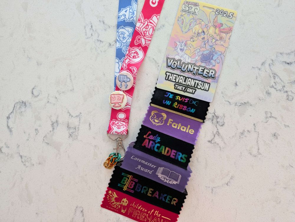 A volunteer badge from SGDQ 2025. It has the username "theValiantSun" and pronouns "they/any". Attached to it are 6 ribbons: a ribbon that says "je suis un ribbon", Frame Fatales, Lady Arcader's, Loremaster Award, Binary Breakers, and a "children of the Firehawk" ribbon. The event lanyard is also displayed with a badge for stream tech and setup on it. 