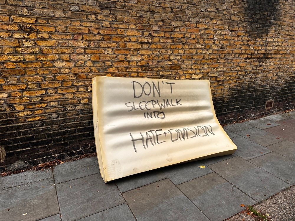 An old mattress lying on a street with the words ‘DON’T SLEEPWALK INTO HATE AND DIVISION’ on it.