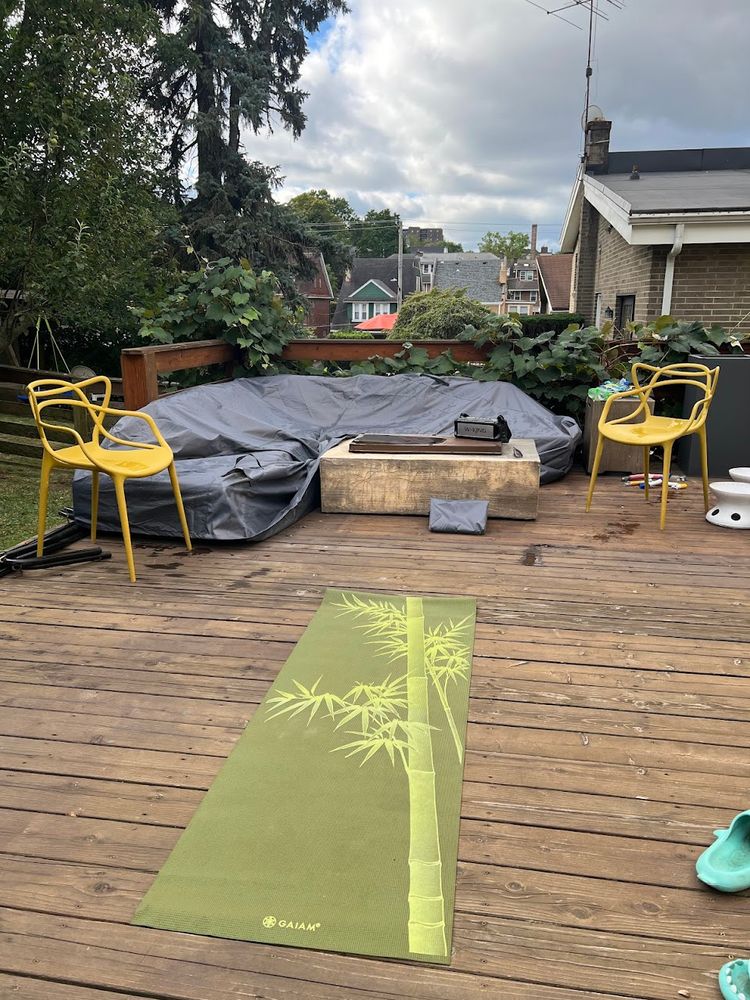 Wooden deck with a yoga mat stretched across it. The view behind is over a propane fire pit with blutooth speaker and houses downhill in the distance.