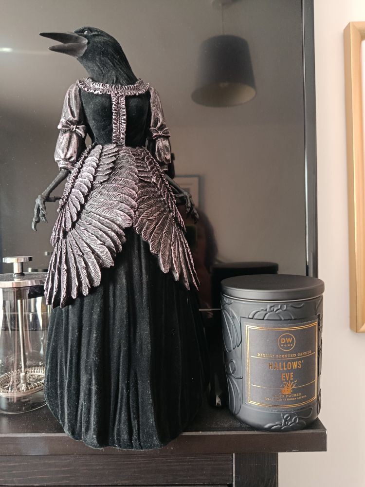 A statue of an anthropomorphic crow dressed in long black velvet dress embellished with feathers and ruffles, standing next to a black glass jar candle with pumpkins carved into the glass