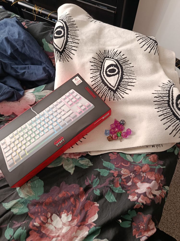 A boxed white mechanical gaming keyboard with rainbow backlights, a white woollen blanket and an assorted handful of coloured dice on a black floral bedspread.