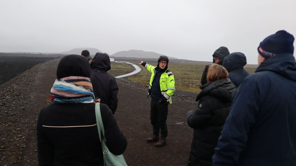 a man in a yellow vest pointing towards a road and a mountain. People around him, standing on top of a gravel barrier