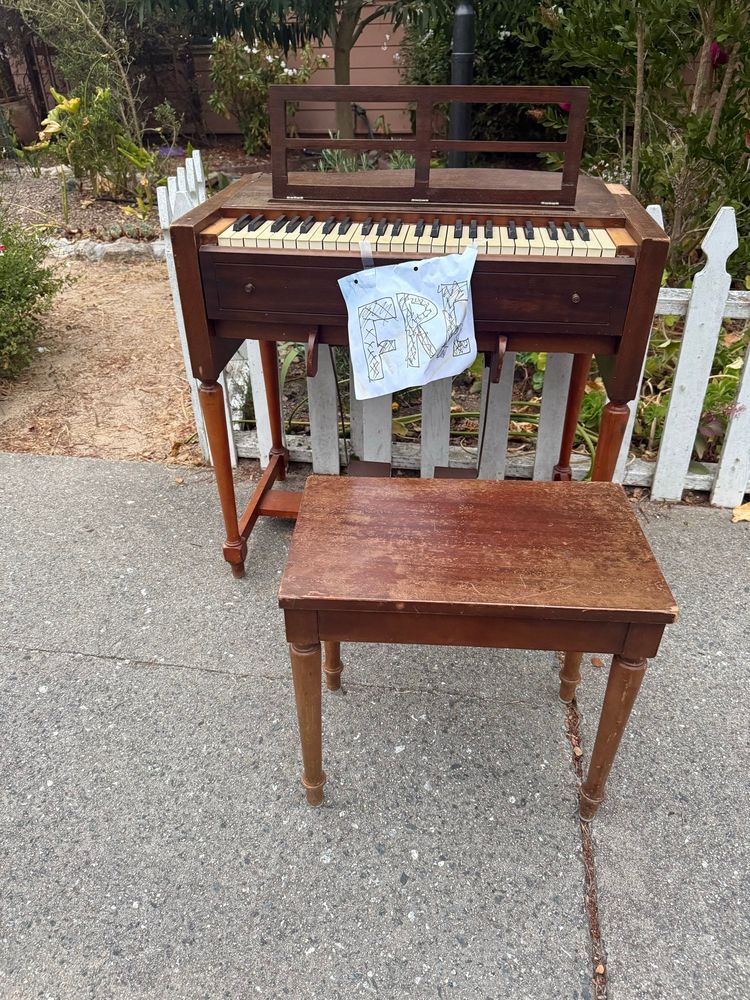 Antique reed organ and stool with “FREE” sign on sidewalk in front of a garden and a white picket fence.