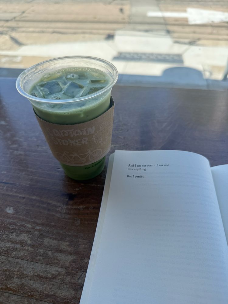 The Sky Isn’t Blue by Janice Lee paperback (Civil Coping Mechanisms, 2016) on table/bar at Captain + Stoker Coffee Roasters in Monterey, CA. View of street through window. Iced matcha latte (with oat milk) to the left of book.