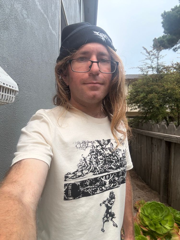Selfie with back of house, succulents, dirt area behind house, wooden fence, trees, adjacent apartment complex, and overcast sky in background. I’m wearing a Late Music shirt and Fire-Toolz beanie.