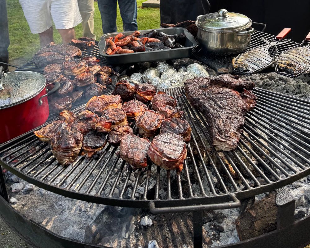 A large round bbq fueled by coals below stocked with many cuts of beef, sausages, jacket potatoes, and whole fish. 