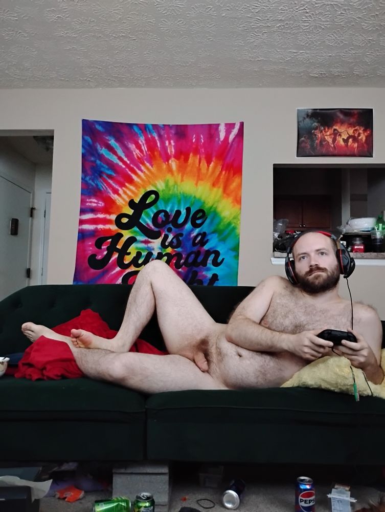 Reunan lays naked on his futon while playing on his Xbox and wearing a chat headset. The tie dye love is a human right tapestry can be seen in the background, a few empty pop cans can be seen on the floor, and the dancing naked women painting can be seen on the wall behind him.