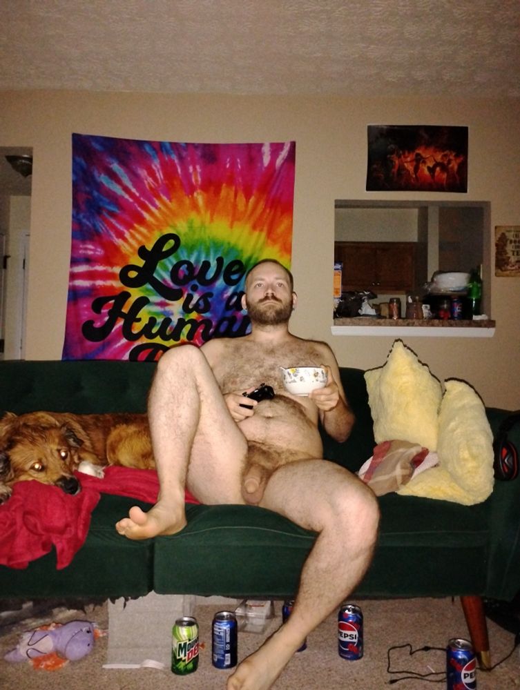 Reunan sits naked on his futon, holding a bowl of cereal (frosted flakes) and his Xbox controller. A few pop cans can be seen on the carpet beneath the futon. His puppy has decided to lay on the futon next to him.