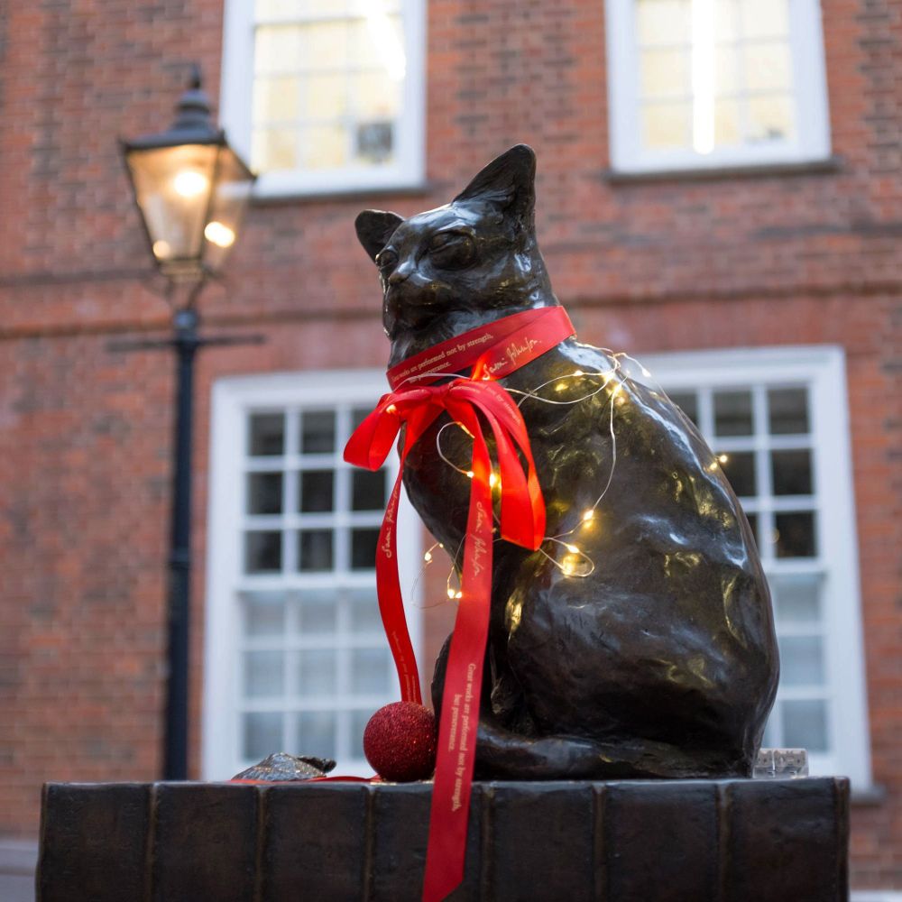 Bronze statue of Hodge the cat outside Dr Johnson’s House, decorated for Christmas with a red ribbon around its neck, fairy lights, and a red bauble. The brick courtyard and windows of the historic house are visible in the background.