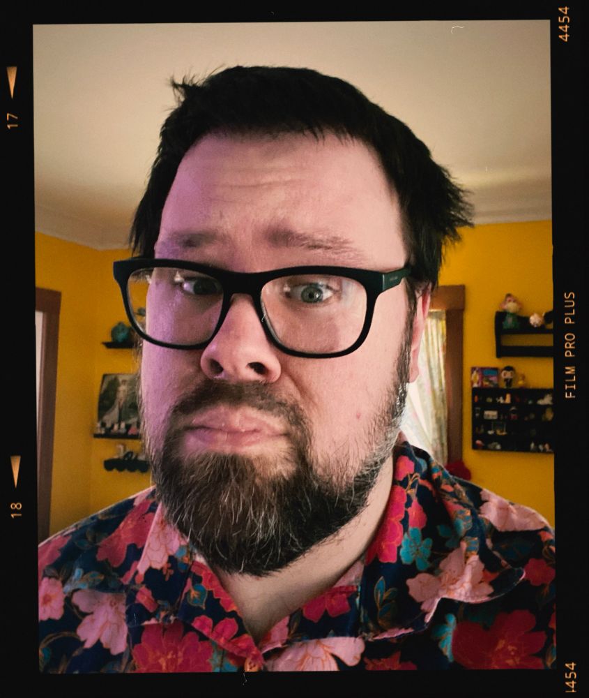 A selfie of me. A man with glasses and full beard. wearing a floral pink, red and blue shirt in front of a yellow wall with a worried look on their face and disheveled hair. Sorry for the run-on sentence.
