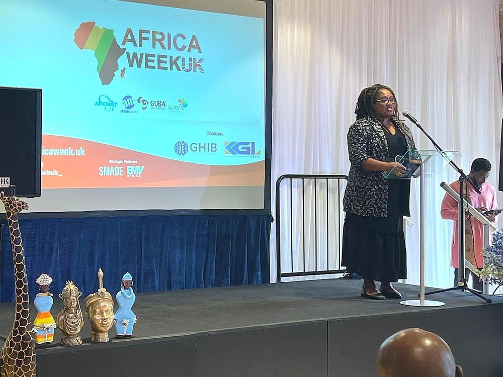Bell Ribeiro-Addy MP speaks at a podium during Africa Week UK, with a colorful presentation displayed on a screen behind her. Traditional African statues are visible on the left.