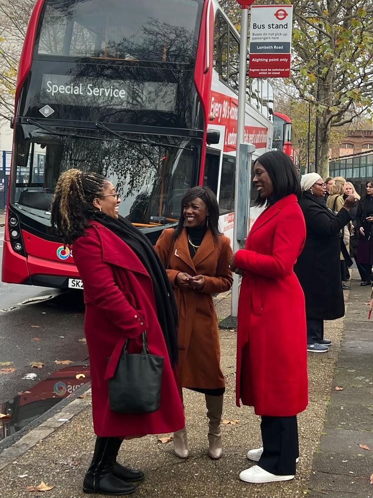 Three individuals stand and chat on a sidewalk beside a red double-decker bus labeled “Special Service.” Two wear red coats, one wears a brown coat. A bus stop sign reads “Bus stand – Lambeth Road – Adjoining point only.” Fallen leaves and tree-lined streets suggest it is autumn.