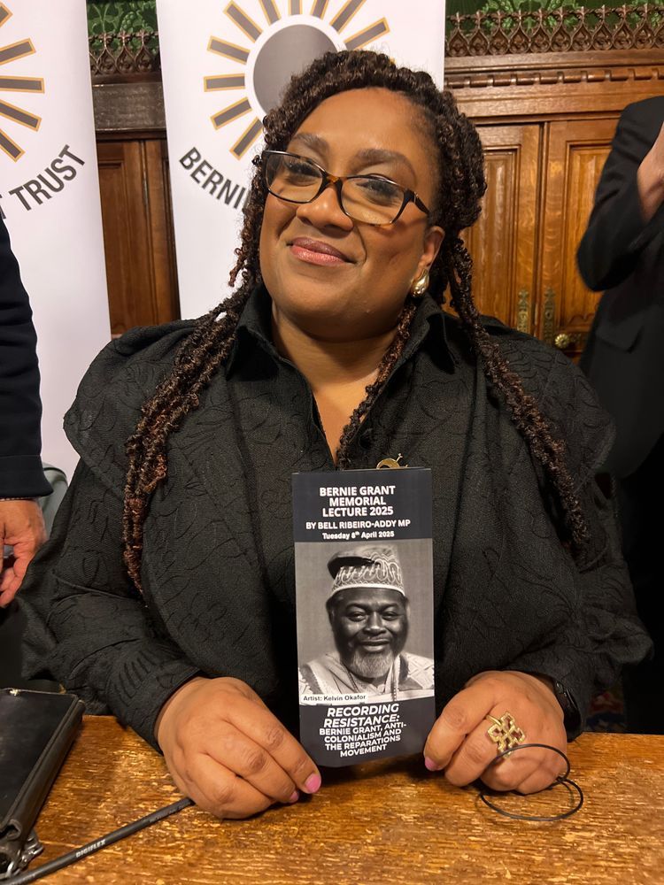 Bell smiles, sitting at a table in a parliamentary committee room, holding a programme for the Bernie Grant Memorial Lecture 2025. Behind her: a banner for the Bernie Grant Trust.