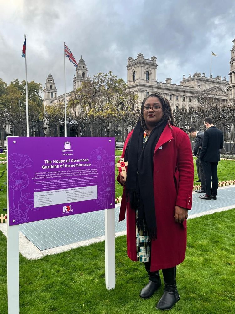 Bell stands in the House of Commons Gardens of Remembrance, holding a small wooden remembrance cross. She is wearing a long red coat, black scarf, and black boots. Behind her is a purple information sign about the Gardens of Remembrance, green grass with small memorial markers, and historic Whitehall buildings with flags flying on tall flagpoles under a cloudy sky.