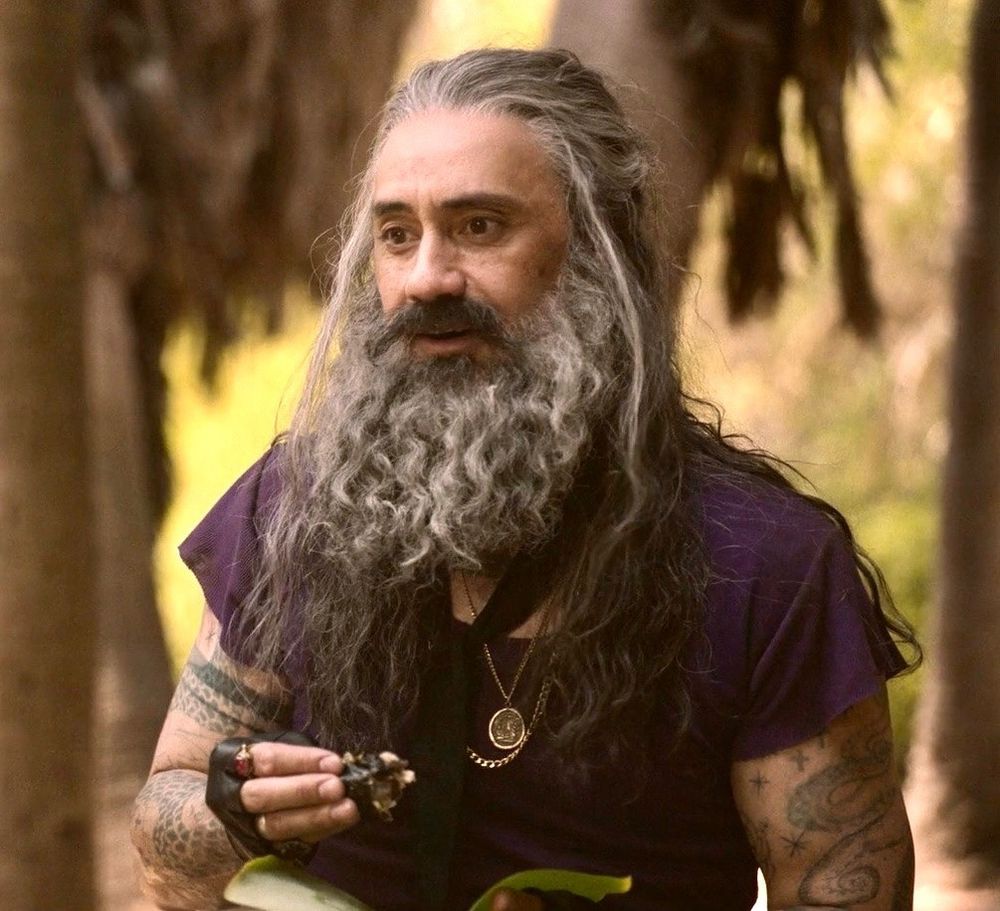 Beautiful, bearded Ed Teach eating a piece of snake.  He looks happy and thoughtful and is wearing a purple shirt.