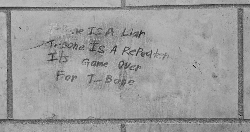 Black and white photo of a building facade with scrawled and half rubbed out inscription: T-Bone is a liar T-Bone is a reapeater Its game over For T-Bone Photo and edit by Shade