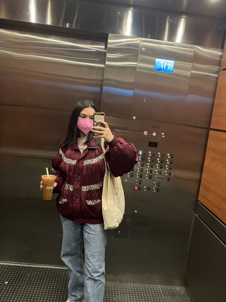 mirror selfie in an elevator, a white woman with a pink mask, jeans, and a burgundy jacket holding an iced coffee and her phone with a tote bag on her shoulder 