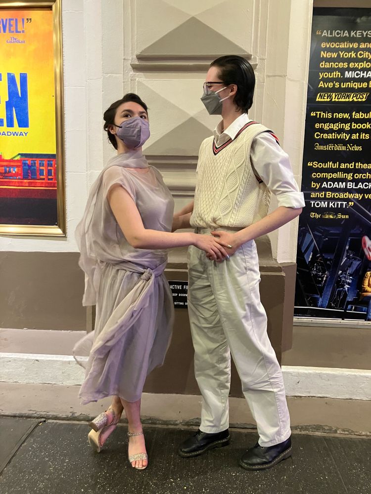 two people holding hands, one looking away. one is in a light purple dress, the other in a white vest, button up, and pants 