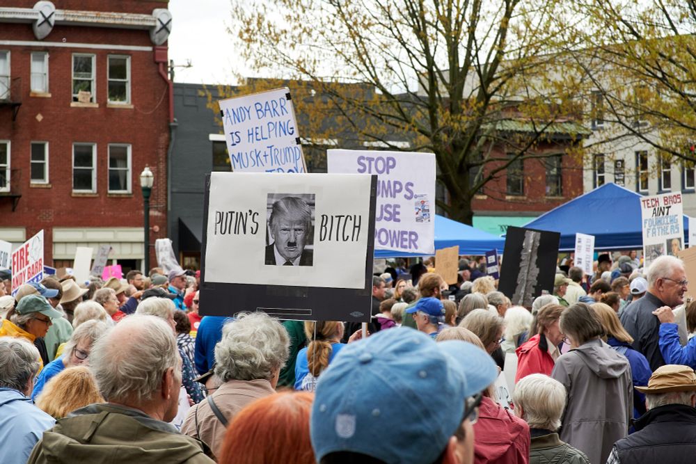 Protestors in Lexington, Kentucky. A prominent sign reads "PUTIN'S BITCH," the two words flanking a photo of Donald Trump wearing a Hitler moustache.