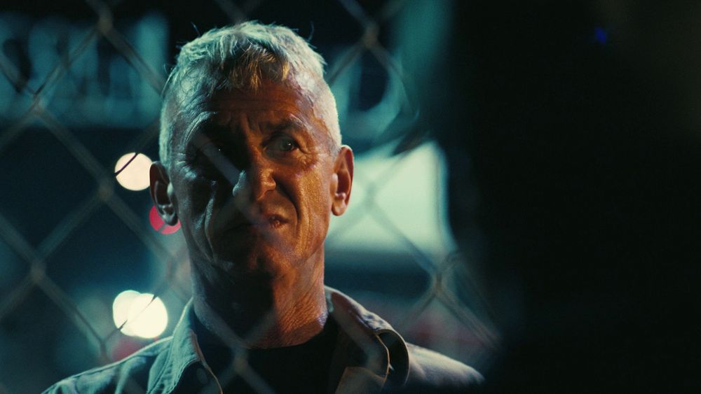 A severe man with hollow eyes stares at figure beyond chainwire fencing. His face is gaunt and bereft of bodyfat and shorn of anything beyond necessity; a tuft of a fringe solely for the minimum viable utility to his unnerving countenance.