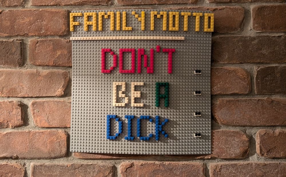 A Lego sign hanging on a red brick wall. The Lego reads - Family Motto -Don't be a dick 