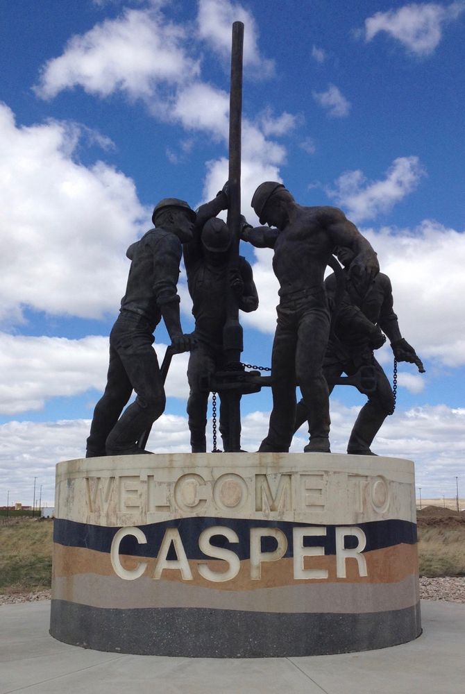 A round pedestal with representative rock layers is inscribed “Welcome to Casper”. Atop sits a bronze statue titled “Man Made Energy” which depicts four oilfield workers, one shirtless with bulging muscles, securing an extension onto a drill head.
