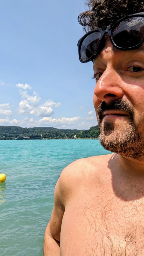 Eine Person namens mauszfabrick blickt in die Kamera, dahinter ist der hellblaue Wörthersee im Strandbad Maria Loretto und eine gelbe Boje zu sehen, mauszfabrick ist gerade bis zur zweiten gelben Boje geschwommen, was an den Wassertropfen auf den Schultern zu erkennen ist