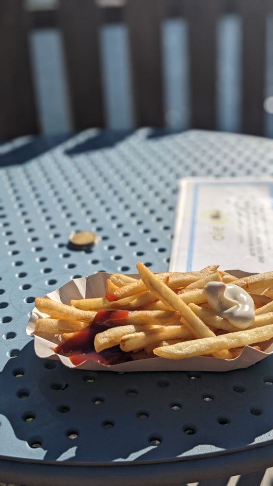 ein Portion Stützfritten auf einem hellblauen See-Badeanstaltskiosktischchen, im Hintergrund ist unscharf ein See zu erkennen 