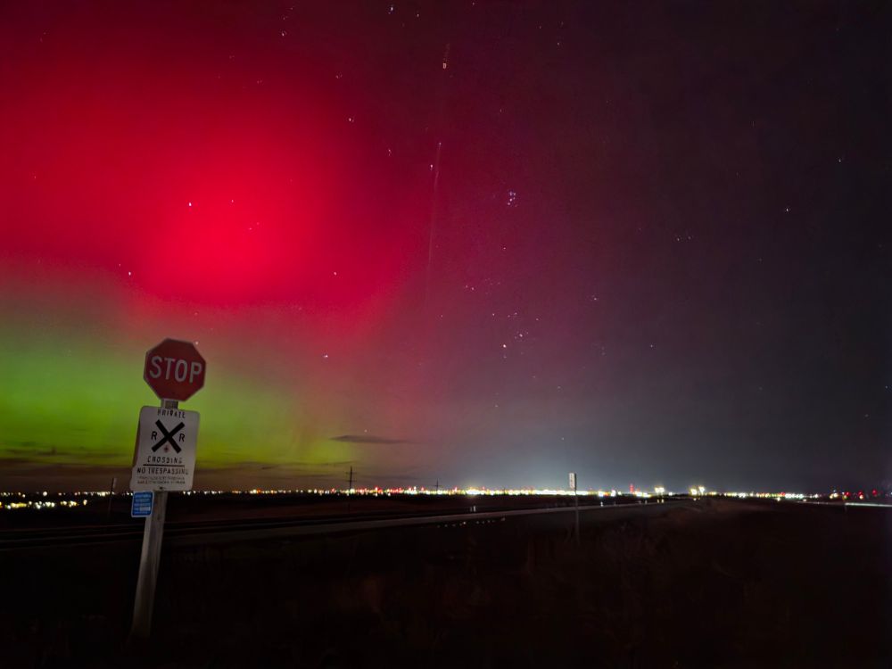 Bright reds sit on top of a band of green. A railroad crossing stop sign is in the foreground. City lights shine on the horizon. 